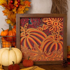 May include: A framed, layered paper art piece depicting pumpkins and autumn leaves in orange, red, and gold hues. A white pumpkin, small orange pumpkins, and a wire basket of pumpkins are in the foreground. A fall wreath is in the background.