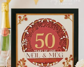 May include: A framed, layered paper art piece celebrating a 50th wedding anniversary. The design features a red circular centerpiece with the number "50" in gold, surrounded by floral cutouts. The names "NEIL & MEG" and the date "January 20, 1968" are also displayed.