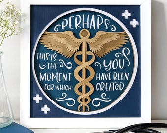 May include: A framed art piece with the text "Perhaps this is the moment for which you have been created." The artwork features a gold caduceus symbol with wings, set against a navy blue background. A stethoscope rests in front of the frame.