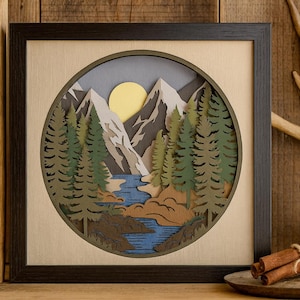 May include: A square framed wall art piece featuring a layered paper cut-out landscape. The scene depicts a blue river flowing through a forest of green pine trees toward snow-capped mountains under a yellow sun. A tag reads My mom & Me.