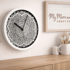 Puede incluir: Un reloj de pared redondo blanco con un diseño de mandala detallado y en capas en tonos grises y negros. Las manecillas del reloj son negras. El reloj está montado en una pared blanca, con un letrero enmarcado que dice "My Mom and Me Craft Co."