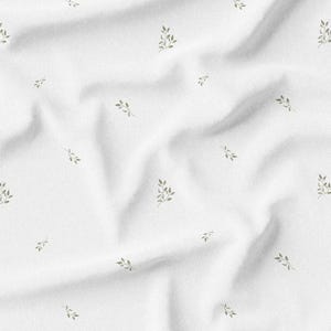 May include: White fabric with a subtle, repeating pattern of small, olive-green sprigs. The fabric is slightly wrinkled, creating shadows and texture. The sprigs are evenly spaced across the surface, offering a delicate, natural aesthetic.