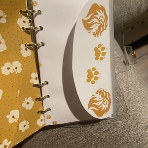 Puede incluir: Una agenda con estampado floral amarillo y blanco con una cubierta de plástico transparente. La agenda tiene anillos dorados y un inserto blanco con un diseño de león y huellas de patas.