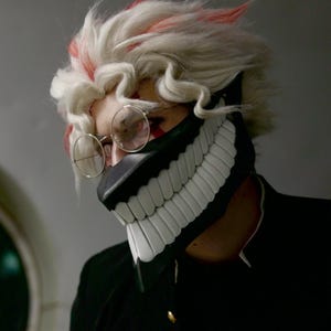 May include: A person wearing a white wig with red streaks and a black and white mask with a large, toothy grin. The mask covers the person's mouth and nose. The person is wearing round glasses.