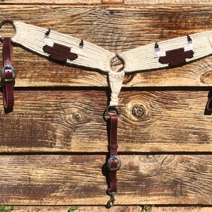 May include: A white and brown woven horse breast collar with brown leather straps and silver buckles. The breast collar has a design of a cow's head with horns.