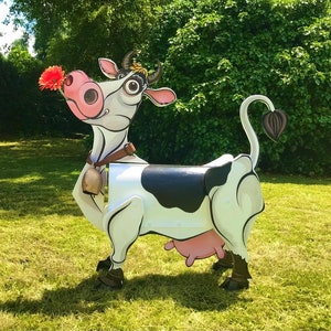 Może przedstawiać: Skrzynka pocztowa w kształcie czarno-białej krowy z różowym wymieniem i dzwonkiem wokół szyi. Krowa patrzy w lewo i ma różowy kwiat w pysku.