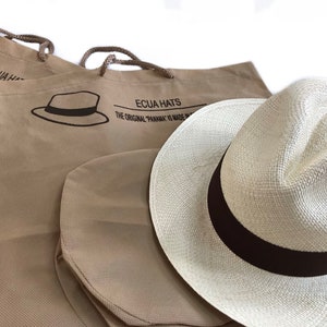 May include: Two brown canvas bags with the text "Ecua Hats The Original "Panama" is Made in Ecuador" printed on them. A white Panama hat with a brown ribbon is sitting on top of the bags.