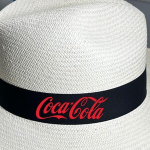 Puede incluir: Un sombrero de paja blanco con una banda negra y el logotipo de Coca-Cola rojo.