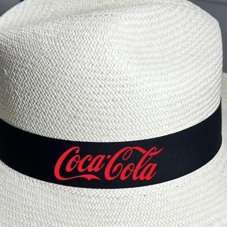 Puede incluir: Un sombrero de paja blanco con una banda negra y el logotipo de Coca-Cola rojo.