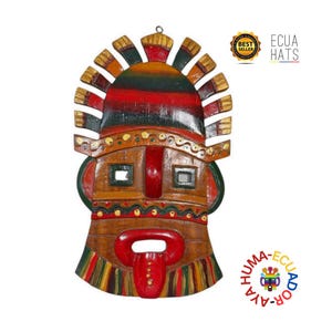 Puede incluir: Máscara de madera tallada a mano con una cara estilizada, con una lengua roja prominente y un tocado con detalles dorados, rojos y verdes. La máscara tiene aberturas cuadradas para los ojos y está pintada en tonos tierra. El texto "ECUA HATS" es visible.