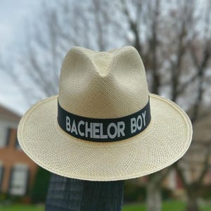 Puede incluir: Un sombrero de paja con una banda negra que dice "Bachelor Boy" en letras blancas.