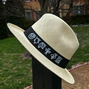 Puede incluir: Un sombrero de paja de color crema con una banda negra que presenta un dise&ntilde;o de una tortuga, hojas, una flor y la palabra "Ecuador".