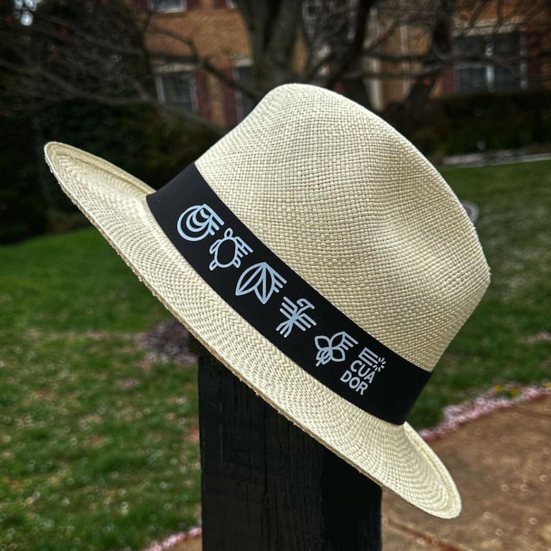 Puede incluir: Un sombrero de paja de color crema con una banda negra que presenta un dise&ntilde;o de una tortuga, hojas, una flor y la palabra "Ecuador".