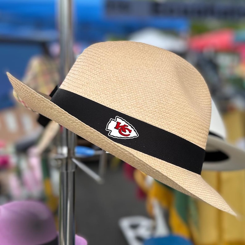 Puede incluir: Un sombrero de paja con una banda negra y un parche con el logotipo de los Kansas City Chiefs en rojo y blanco.