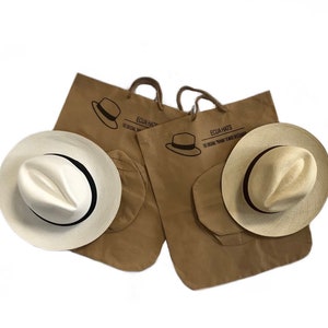 May include: Two brown paper bags with the text "Ecua Hats" and "The Original Panama  Toquilla Straw Hats" printed on them. Each bag contains a white Panama hat with a black ribbon.