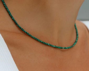 May include: A delicate necklace composed of small, round, green beads. The necklace is worn around the neck, resting against the skin. The beads have a natural, earthy appearance, and the necklace appears to be made of a gemstone.