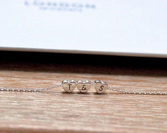 May include: A delicate silver necklace featuring three heart-shaped charms. The charms are engraved with the letters "T", "&", and "S". The necklace is displayed on a wooden surface.
