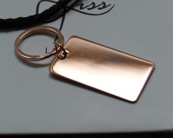 May include: A rose gold rectangular keychain with a matching key ring. The keychain is blank and ready for engraving. A black cord is visible in the background. The word "kiss" is printed on the background.