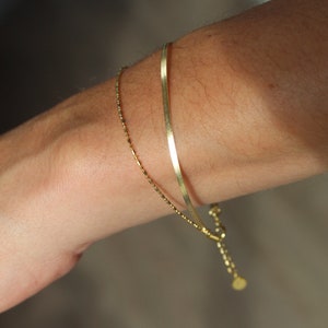 May include: Two gold bracelets on a wrist. One bracelet is a thin chain with small beads, and the other is a thicker, flat chain.
