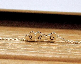 May include: A silver chain necklace with three heart-shaped charms. The charms are engraved with the letters "P", "&", and "G".