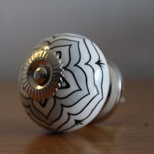 Könnte beinhalten: Ein weiß-schwarzer Möbelknopf mit Muster und einer goldfarbenen Metallbasis. Der Knopf hat eine runde Form mit einem floralen Design. Die Hardware ist für Möbel und Heimdekoration konzipiert.