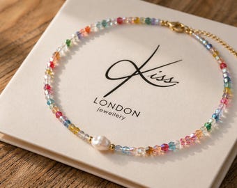 May include: A delicate necklace featuring a strand of colorful, faceted beads in rainbow hues, interspersed with clear beads. A single, lustrous white pearl and gold-toned clasp complete the design. The necklace rests on a white box with the text "Kiss LONDON jewellery".