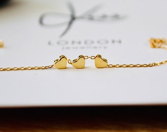 May include: A gold chain necklace with three small heart charms. The necklace is on a white surface with the text "LONDON JEWELRY" visible.