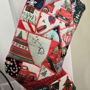 Puede incluir: Un calcetín navideño de patchwork con una variedad de patrones festivos. El diseño incluye camiones rojos, árboles de Navidad, copos de nieve y cajas de regalo. Se ven las palabras "Season's Greetings" y "Home for the Holidays".