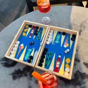 Puede incluir: Un tablero de backgammon de madera con un diseño azul y amarillo que presenta conos de helado, scooters y ramas de olivo. El tablero está abierto y tiene fichas negras y blancas. Dos vasos de cócteles rojos con rodajas de naranja están sobre una mesa gris.