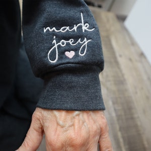 May include: Close-up of a dark gray sweatshirt sleeve with white embroidery that reads "mark joey" with a small pink heart.