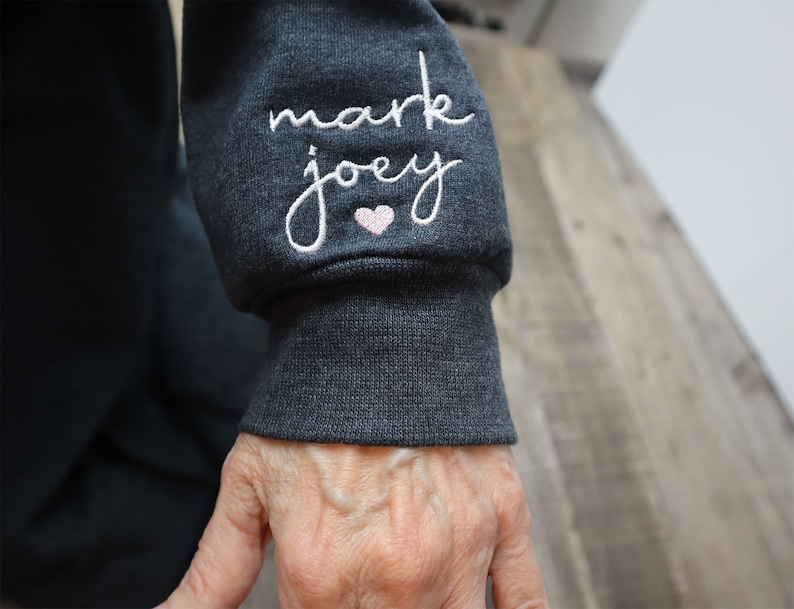 May include: Close-up of a dark gray sweatshirt sleeve with white embroidery that reads "mark joey" with a small pink heart.