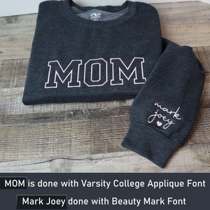 May include: A dark gray long-sleeved shirt with the word "MOM" in white with a pink outline on the front. The shirt has a small heart and the words "mark joey" in pink on the left sleeve.