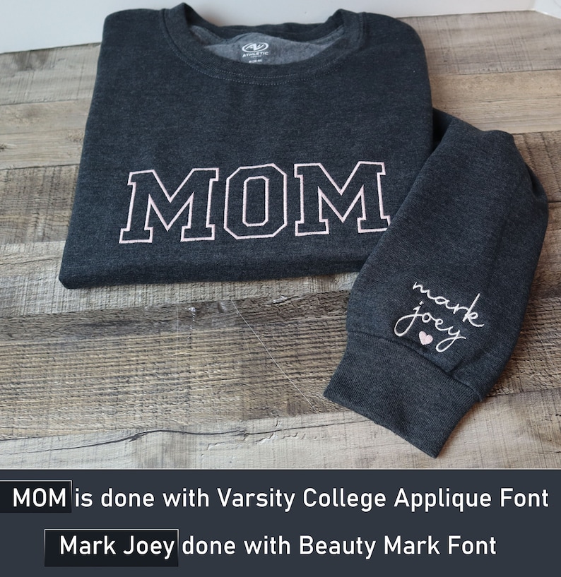 May include: A dark gray long-sleeved shirt with the word "MOM" in white with a pink outline on the front. The shirt has a small heart and the words "mark joey" in pink on the left sleeve.