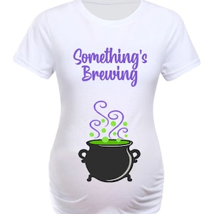 May include: White maternity t-shirt with the phrase "Something's Brewing" in purple script. Below the text is a black cauldron with green bubbling liquid and purple steam. The shirt is designed to accommodate a growing belly.