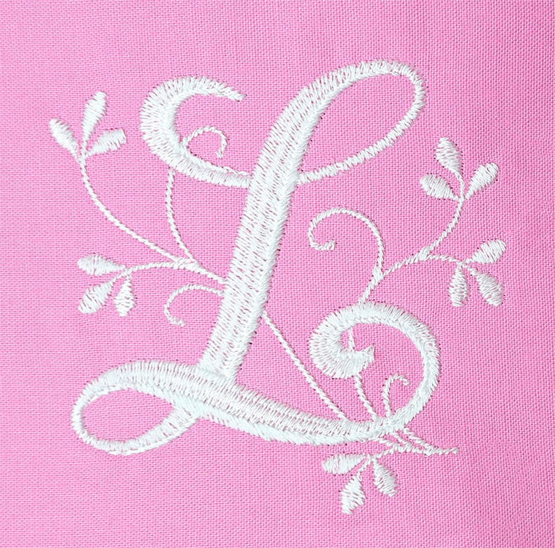 K&ouml;nnte beinhalten: Wei&szlig; gestickter Buchstabe L mit floralen Verzierungen auf einem rosa Stoffhintergrund.