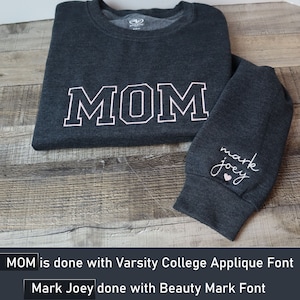 May include: A dark gray long-sleeved shirt with the word "MOM" in white with a pink outline on the front. The shirt has a small heart and the words "mark joey" in pink on the left sleeve.