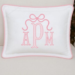 May include: White rectangular pillow with a pink embroidered monogram and bow design. The pillow has a pink border and is placed on a light-colored surface. The monogram features the letters A, P, and M.