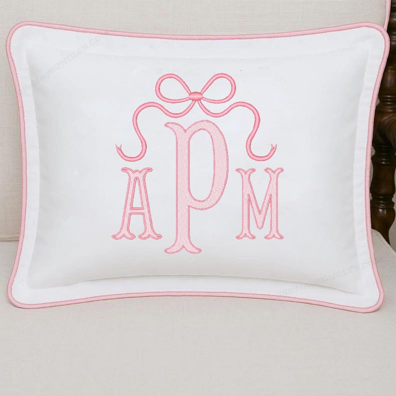May include: White rectangular pillow with a pink embroidered monogram and bow design. The pillow has a pink border and is placed on a light-colored surface. The monogram features the letters A, P, and M.