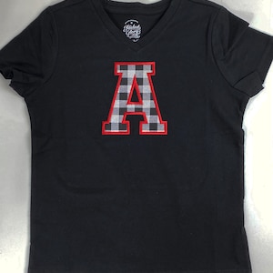 May include: Black v-neck t-shirt with a red and white checkered letter A in a varsity style font.