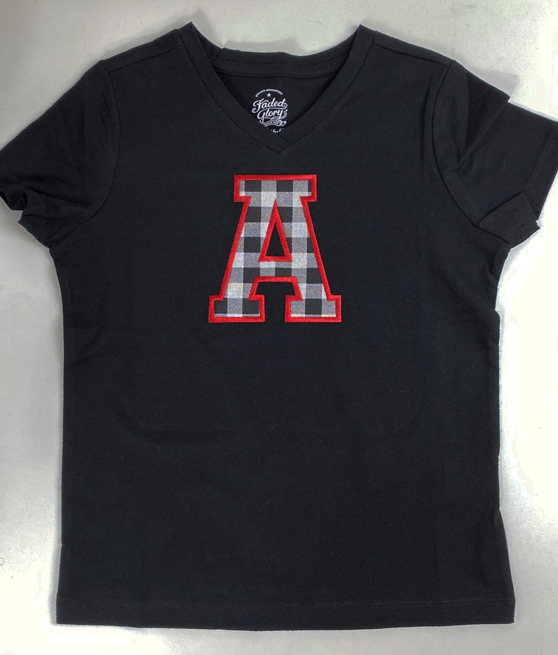 May include: Black v-neck t-shirt with a red and white checkered letter A in a varsity style font.