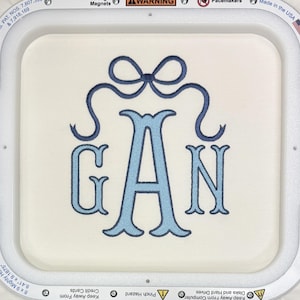 May include: Embroidered monogram featuring the letters G, A, and N in light blue, with a decorative bow at the top. The letters are in a classic serif font, stitched on a cream-colored fabric. The design is framed by a white embroidery hoop.