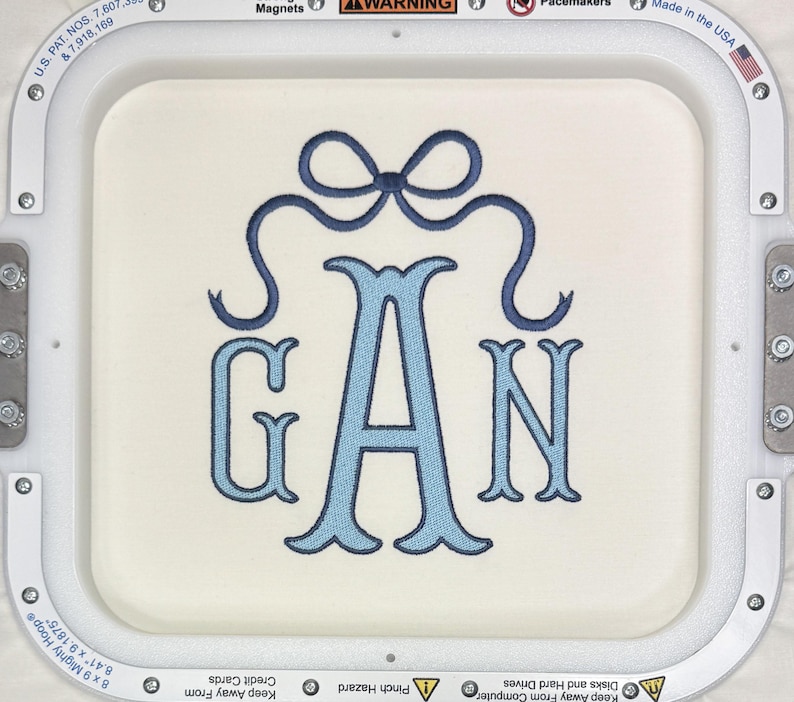 May include: Embroidered monogram featuring the letters G, A, and N in light blue, with a decorative bow at the top. The letters are in a classic serif font, stitched on a cream-colored fabric. The design is framed by a white embroidery hoop.
