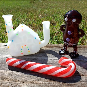 May include: Three glass Christmas-themed smoking pipes. A white gingerbread house pipe, a brown gingerbread man pipe, and a red and white candy cane pipe.