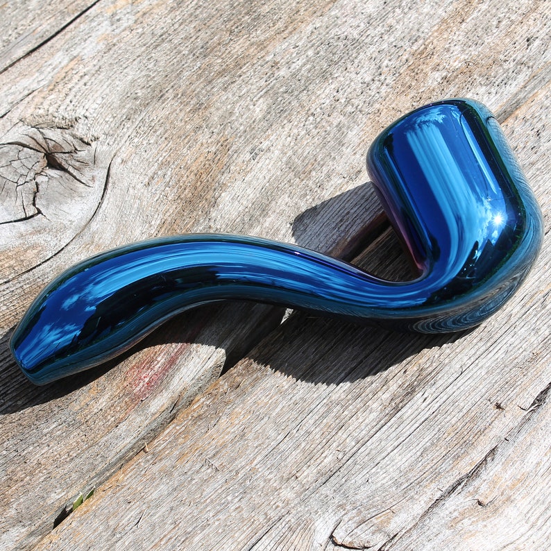 May include: A blue glass pipe with a curved stem and a round bowl. The pipe has a metallic finish and a reflective surface.