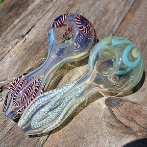 Könnte beinhalten: Zwei gläserne Rauchpfeifen mit aufwendigen Designs. Eine Pfeife hat ein rotes und weißes Spiralmuster, während die andere ein blaues und weißes Spiralmuster hat.