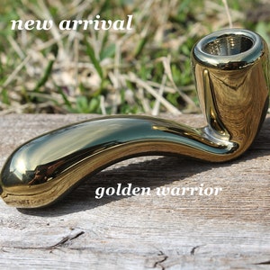 May include: A shiny gold ceramic pipe with a curved stem and a round bowl. The pipe is sitting on a wooden surface. The text "golden warrior" is visible below the pipe.