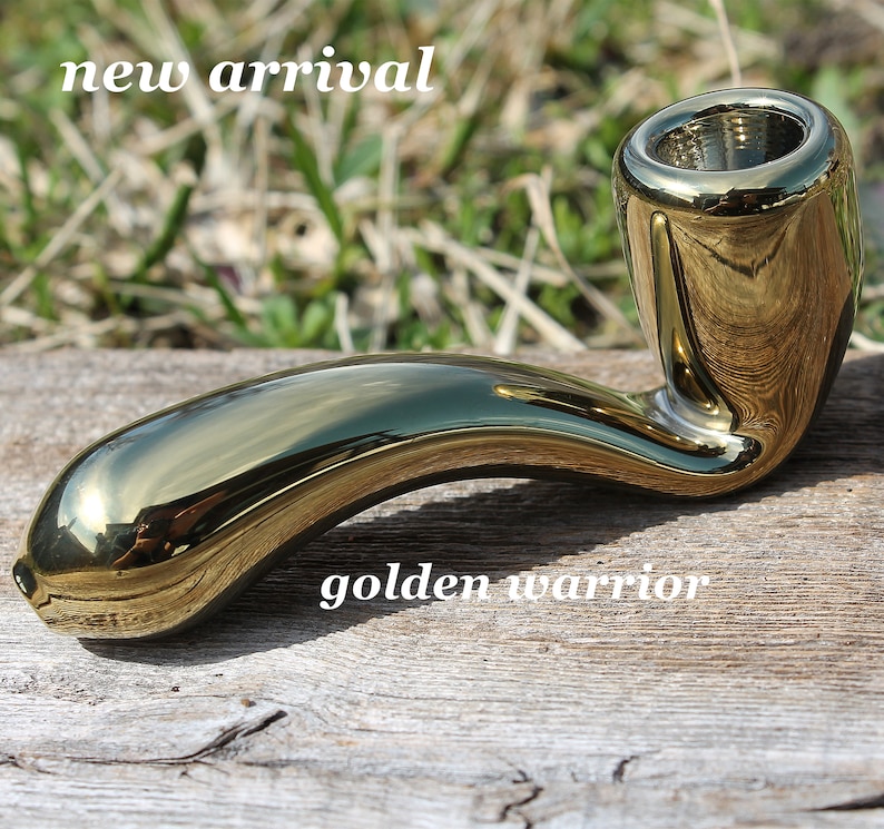 May include: A shiny gold ceramic pipe with a curved stem and a round bowl. The pipe is sitting on a wooden surface. The text "golden warrior" is visible below the pipe.
