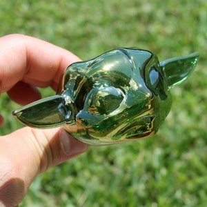 K&ouml;nnte beinhalten: Eine gr&uuml;ne Glasfigur eines Charakters mit gro&szlig;en Ohren und einem breiten Mund. Die Figur ist gl&auml;nzend und hat einen metallischen Schimmer.