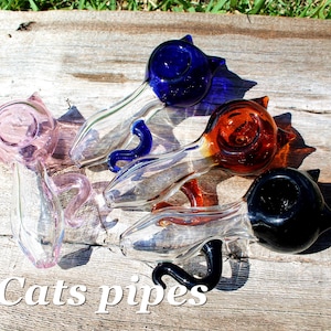 Puede incluir: Cuatro pipas de gato de vidrio en diferentes colores: rosa, azul, naranja y negro. Cada pipa tiene un diseño de cabeza de gato y un cuerpo de vidrio transparente.