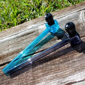 Puede incluir: Dos pipas para fumar de vidrio con cuencos cuadrados y boquillas negras. Las pipas son azules y moradas.
